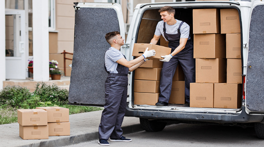 alt= Two workers in uniform load cardboard boxes into the back of a van on a sidewalk, with some boxes already placed inside the van and one on the ground nearby.